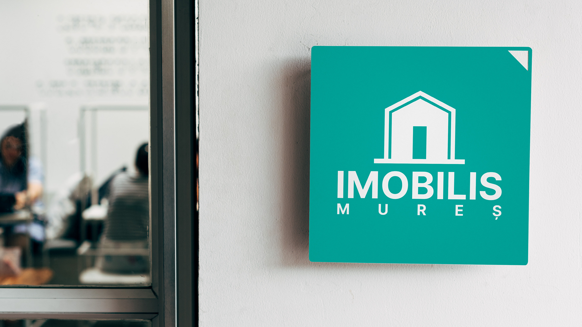 Square turquoise plaque with the white Imobilis Mureș logo, mounted on a white wall beside a glass door