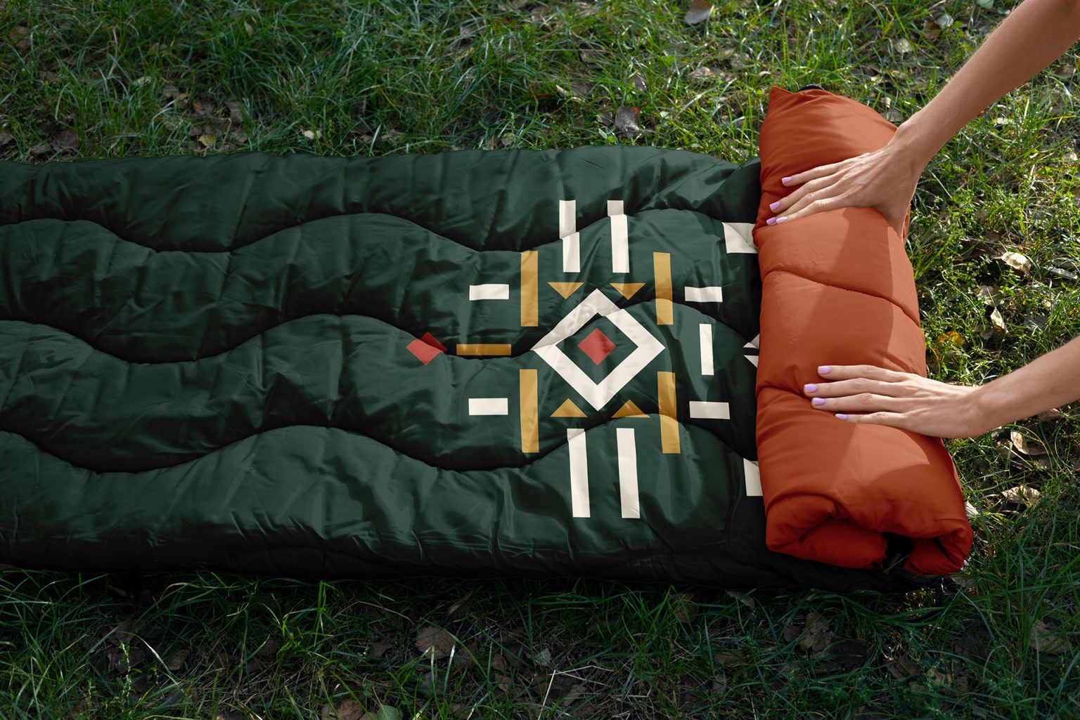 Dark green sleeping bag laid out on grass, with the emblem's central motif in white, ochre and red
